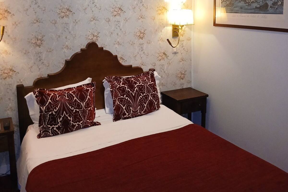 Una habitación de hotel con una cama, papel tapiz floral, un cuadro en la pared y una mesita de noche.
