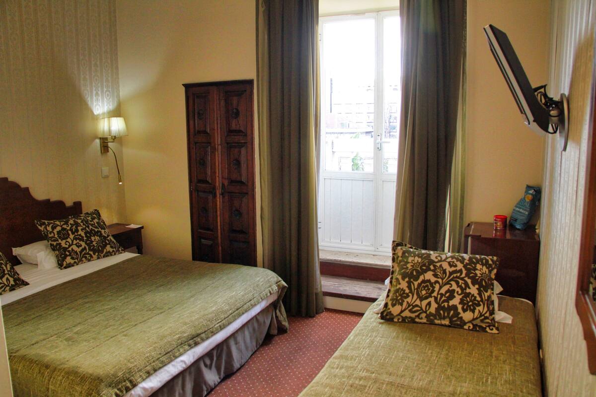 Habitación de hotel con dos camas, un armario de madera, una ventana con cortinas, un televisor montado en la pared y una mesita de noche con lámpara.