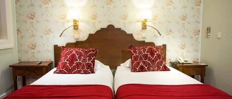A hotel room with two beds, each with a red blanket and matching pillows, a wooden headboard, and a floral wallpaper.