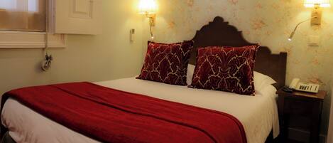 A hotel room with a bed, two wall-mounted lamps, a telephone, and a window with blinds.