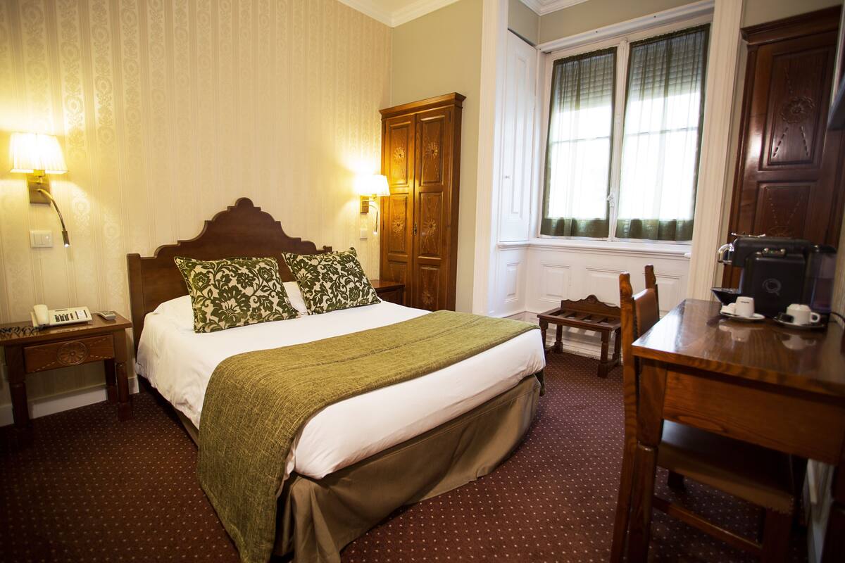Una habitación de hotel con una cama grande, dos lámparas de noche, un armario de madera, una mesita pequeña, una silla y una ventana con cortinas.