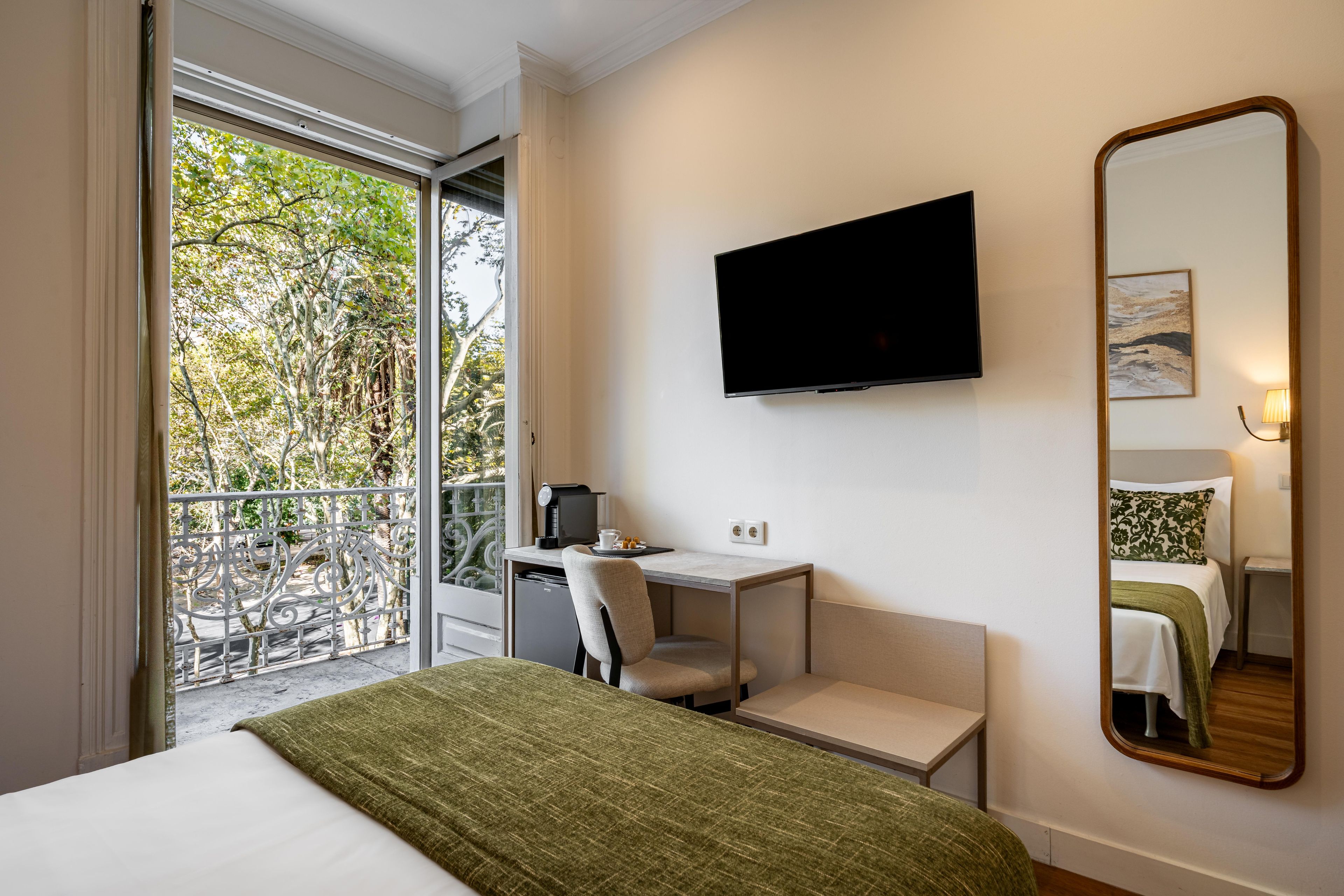 A hotel room with a bed, a desk, a chair, a television, and a balcony with a view of trees.