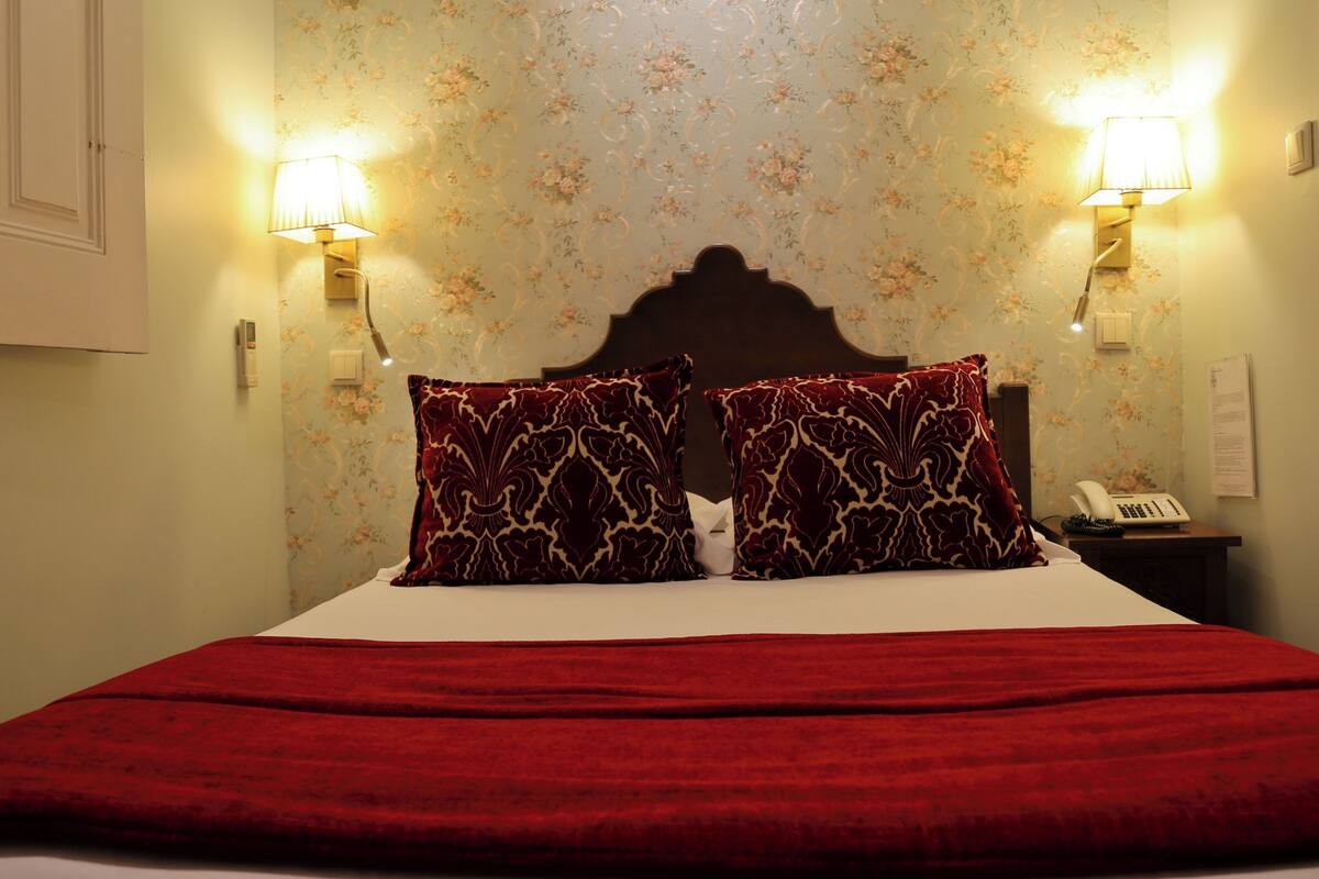 Una habitación de hotel con cama, dos lámparas de pared, un teléfono y papel tapiz floral.