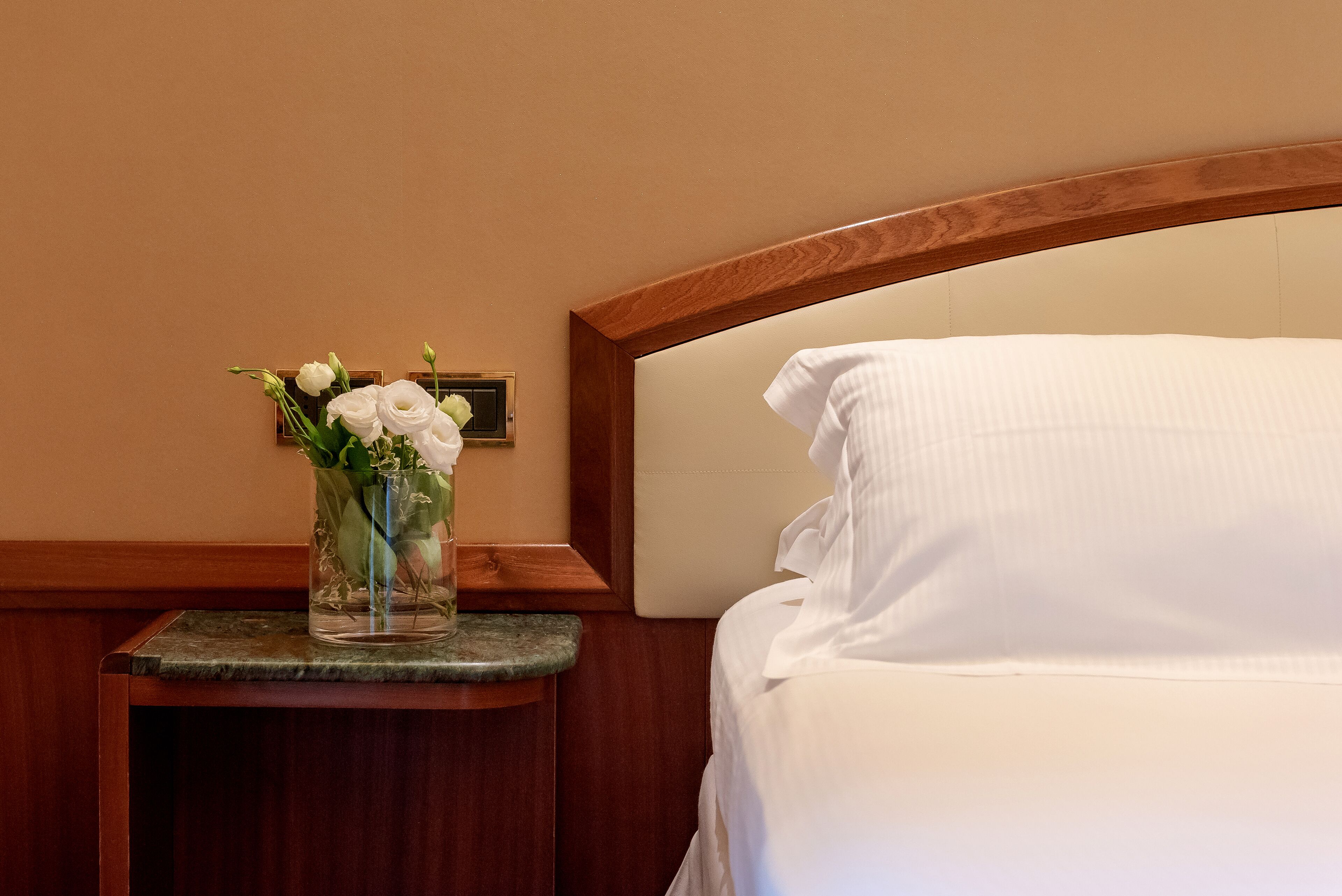 Una cama bien hecha con sábanas blancas, un cabecero de madera y una pequeña mesa con un jarrón de flores blancas.