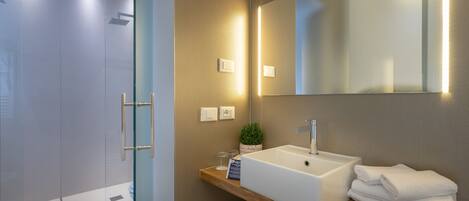 A modern bathroom with a glass shower enclosure, a white sink with a mirror above, and a wooden shelf with towels.