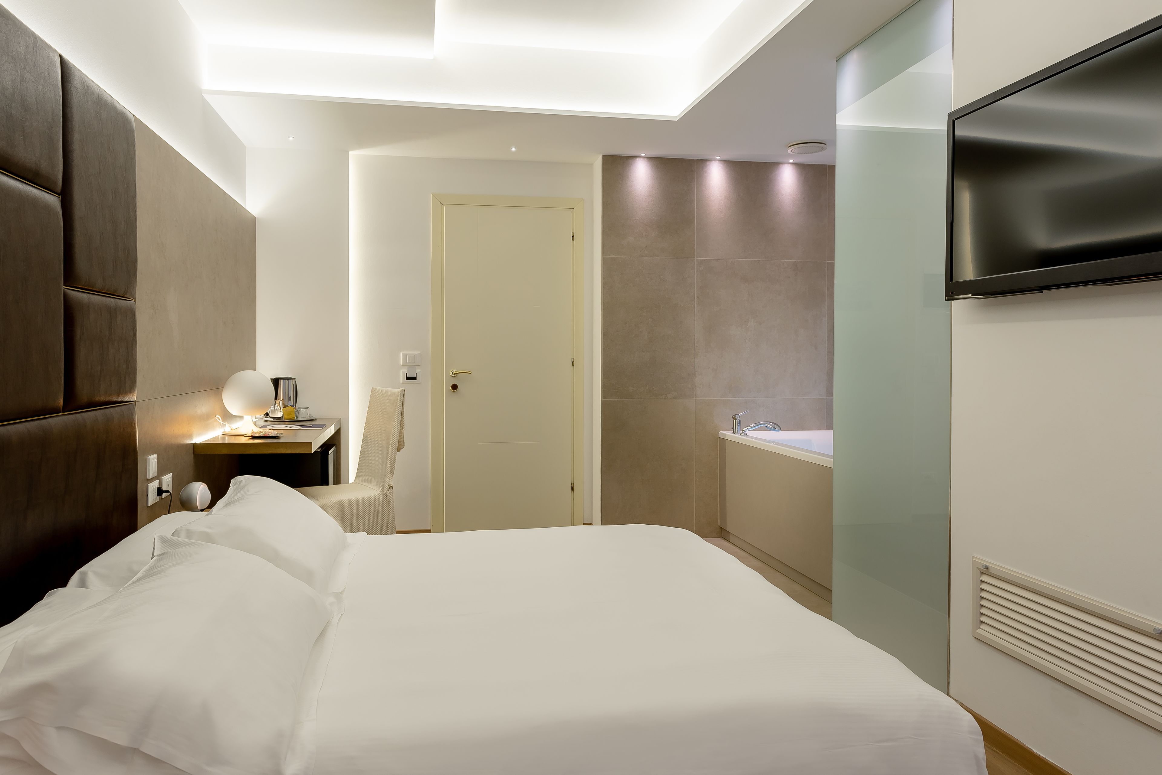 Una habitación de hotel moderna con una cama grande, una mesita de noche con lámpara, un televisor montado en la pared y un baño con bañera.