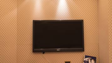 A hotel room with a flat-screen TV mounted on the wall, a wooden desk with a chair, and a small cabinet below.