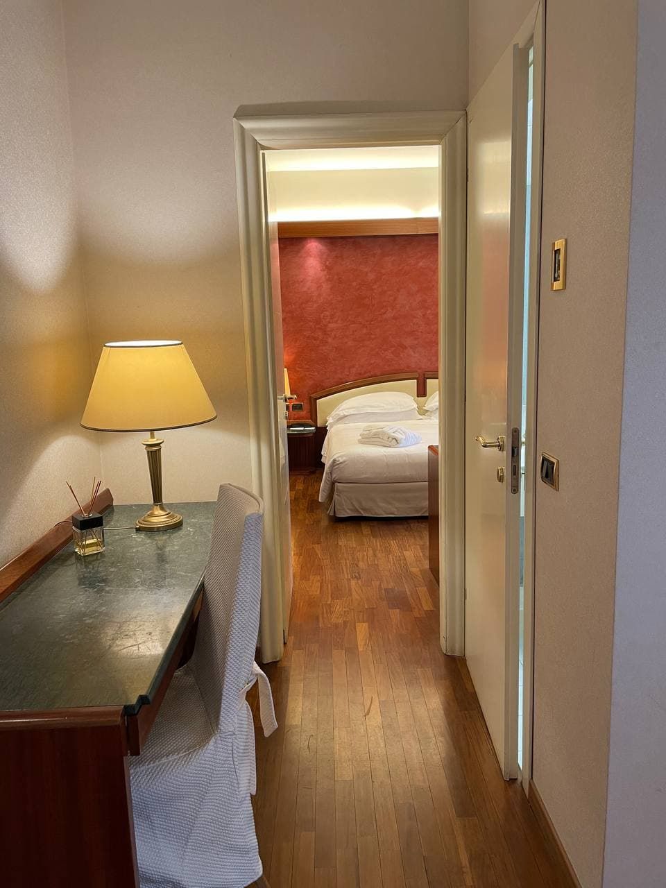 Quarto de hotel com piso de madeira, uma escrivaninha com luminária, uma cadeira, uma cama com roupa de cama branca e uma parede com detalhes em vermelho.