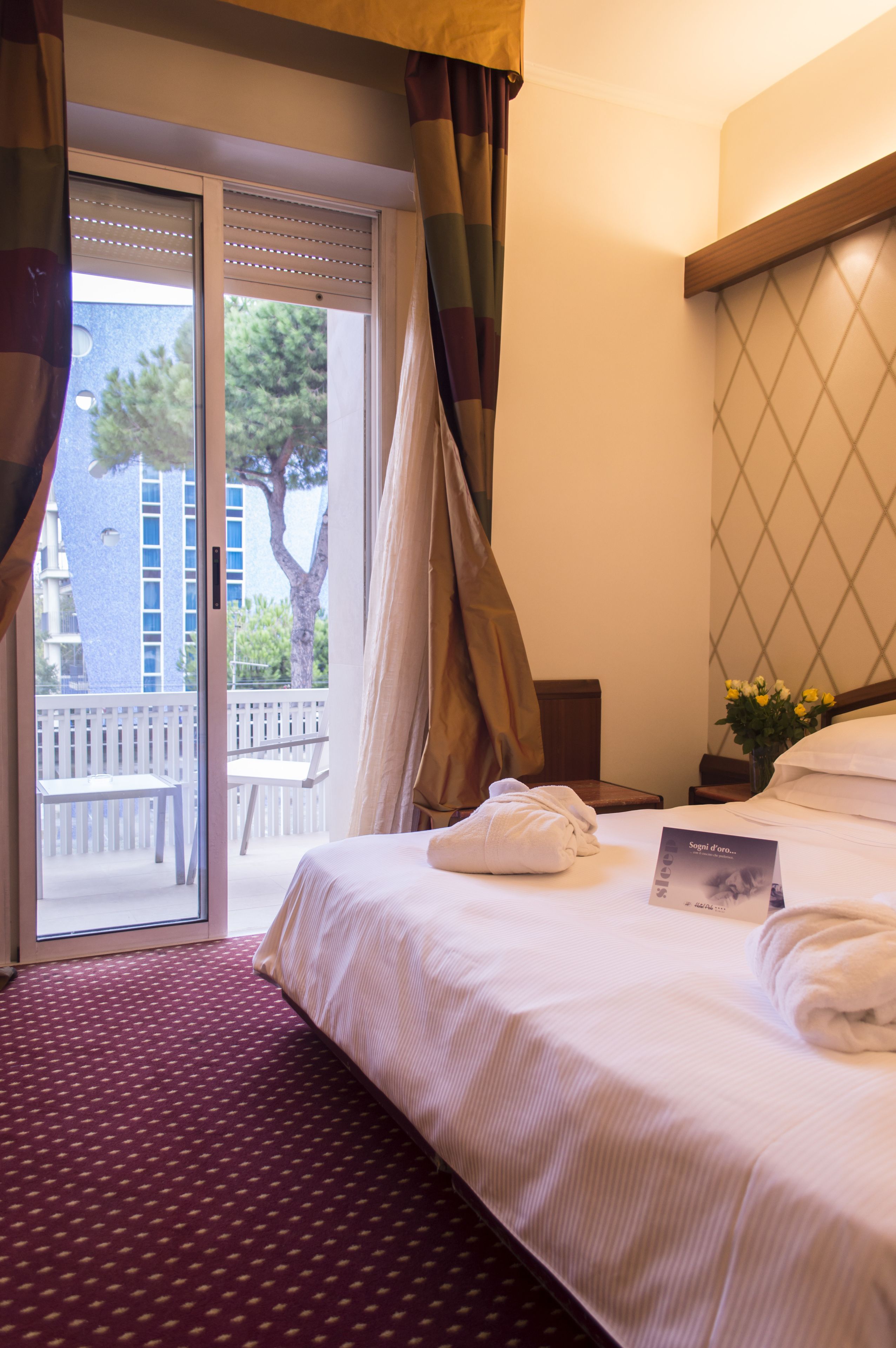 A hotel room with a bed, a nightstand, a window with curtains, a view of the outside, and a small balcony with a chair.