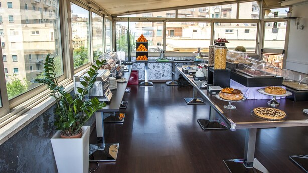 A modern dining area with a long counter, multiple seating options, and a view of the city through large windows. - Hotel Delle Province (Rome)