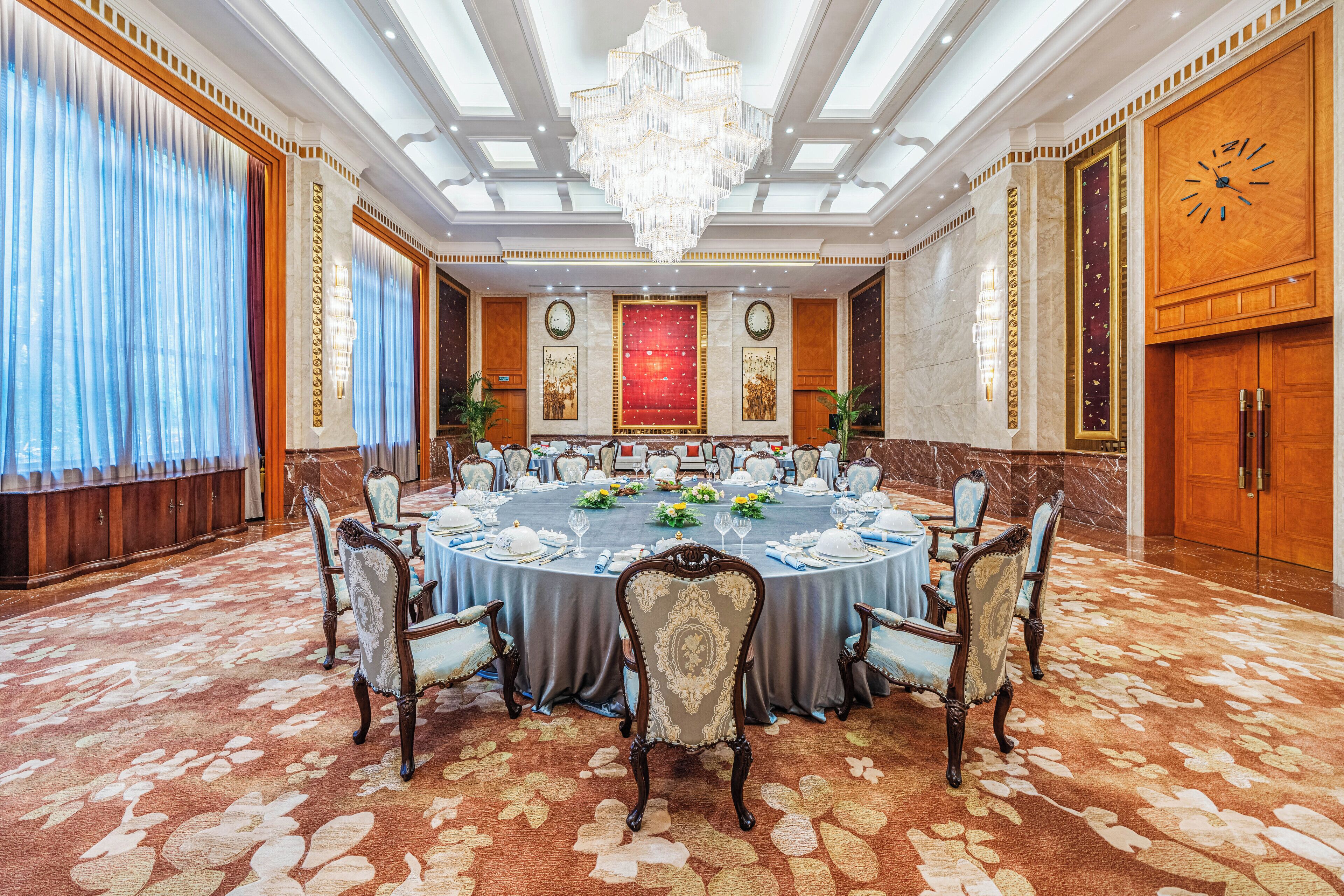 Un comedor formal con una mesa redonda dispuesta para un banquete, rodeada de sillas ornamentadas, una araña y un gran reloj en la pared.