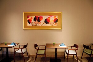 A dining area with tables and chairs, a framed painting on the wall, and a small plant on each table.