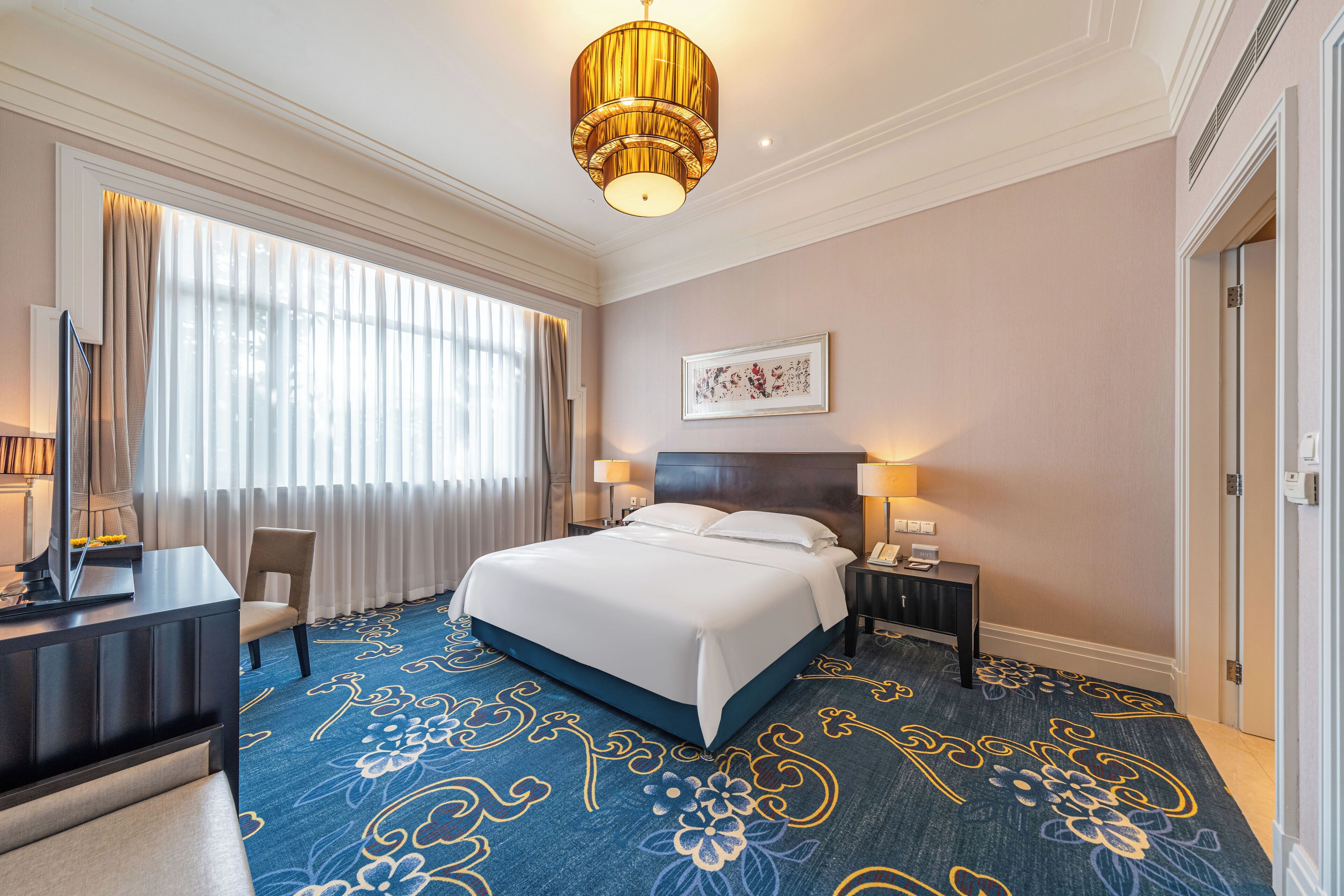 A hotel room with a large bed, a desk, a chair, a nightstand, and a wall-mounted lamp.