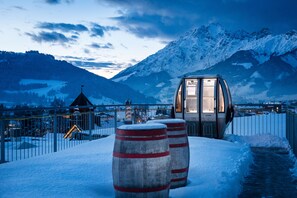 A snow-covered outdoor area with a glass-enclosed structure, possibly a viewing platform or small building, and two large barrels. - Hotel die Hindenburg (Saalfelden am Steinernen Meer)