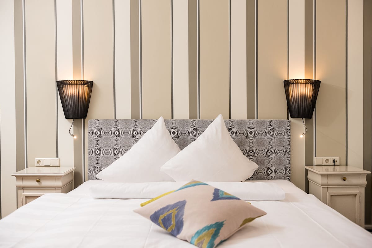 A neatly made bed with white linens and decorative pillows, a grey patterned headboard, and two wall-mounted lamps on either side.