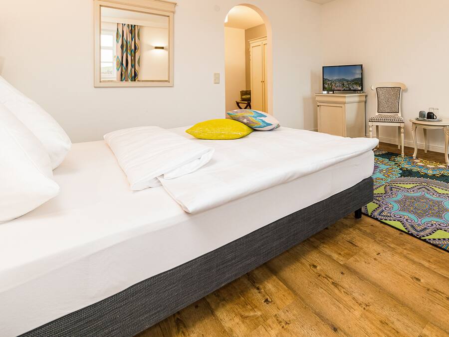 A neatly made bed with white linens and a gray headboard, a small table with a coffee pot, a television on a stand, and a patterned rug on the floor.