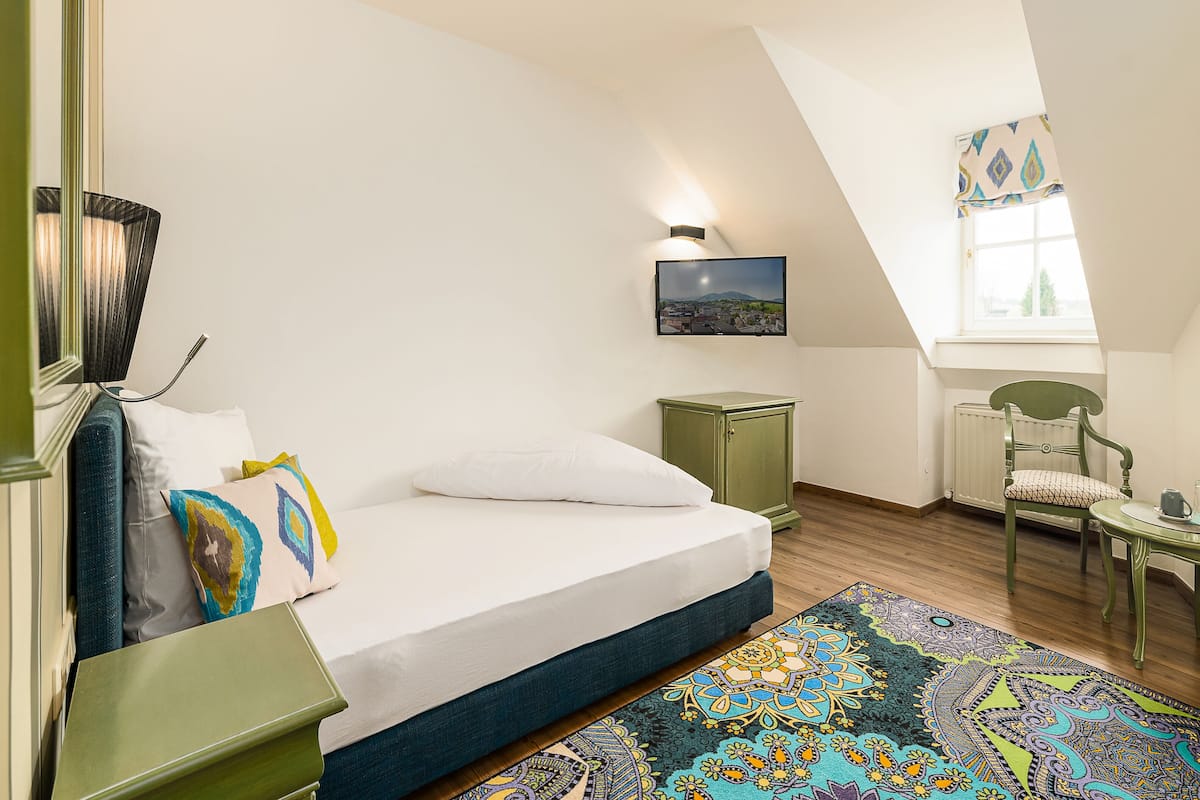 A bedroom with a bed, a green cabinet, a chair, a small table, and a window with a view of a cityscape.