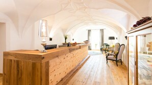 A wooden reception desk with a modern lamp, a vase with flowers, and a fruit bowl. There is a dining area with chairs and a table in the background. - Hotel die Hindenburg (Saalfelden am Steinernen Meer)