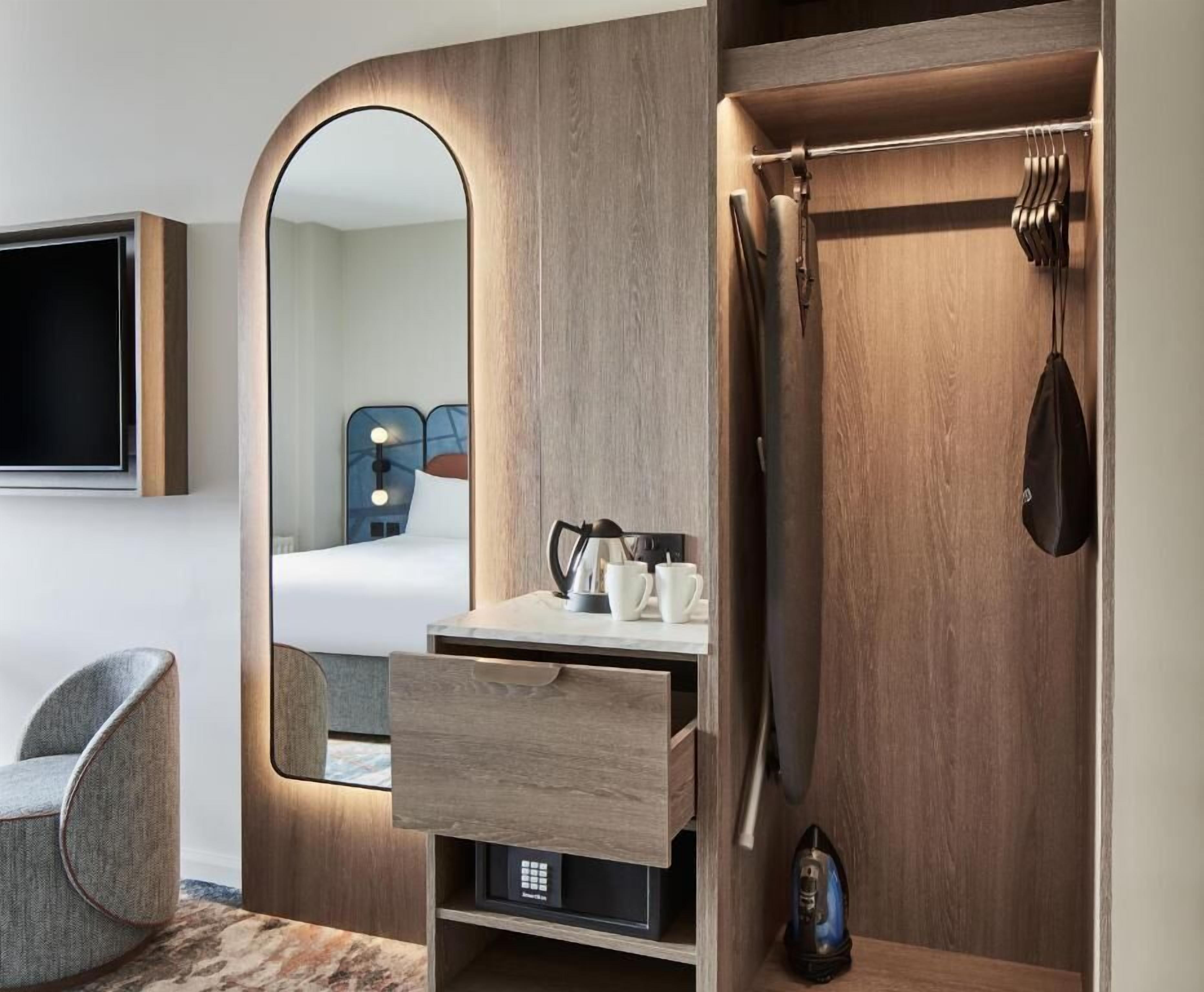 Habitación de hotel moderna con tocador de madera, espejo, televisión de pantalla plana, una silla y armario con ropa colgada.