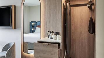 Habitación de hotel moderna con tocador de madera, espejo, televisión de pantalla plana, una silla y armario con ropa colgada.