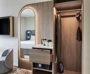 A modern hotel room with a wooden vanity, a mirror, a flat-screen TV, a chair, and a wardrobe with hanging items. - Leonardo Hotel Newcastle (Newcastle-upon-Tyne)