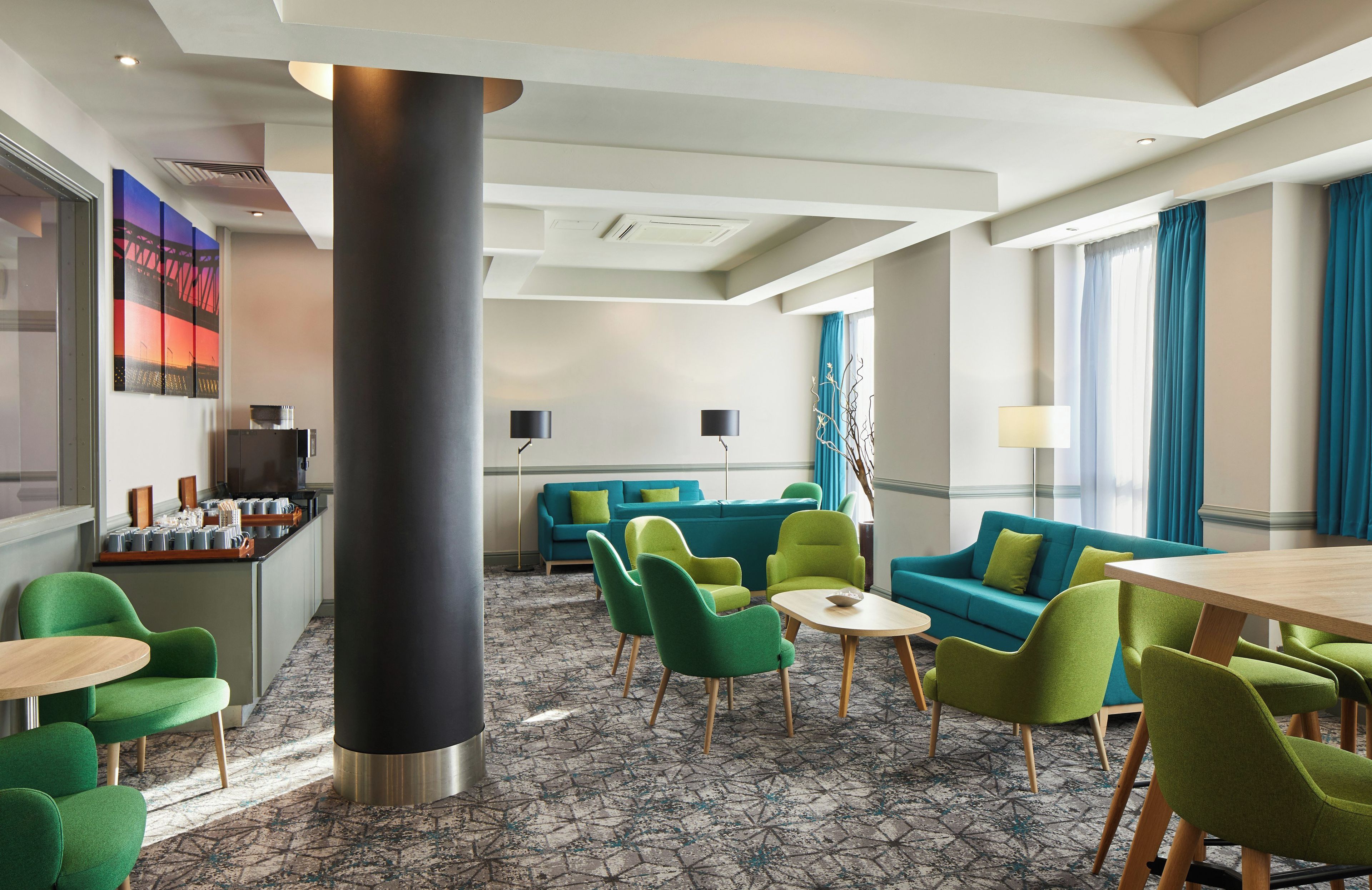 A hotel lobby with a modern design, featuring a large black pillar, green and blue seating, and a coffee bar.