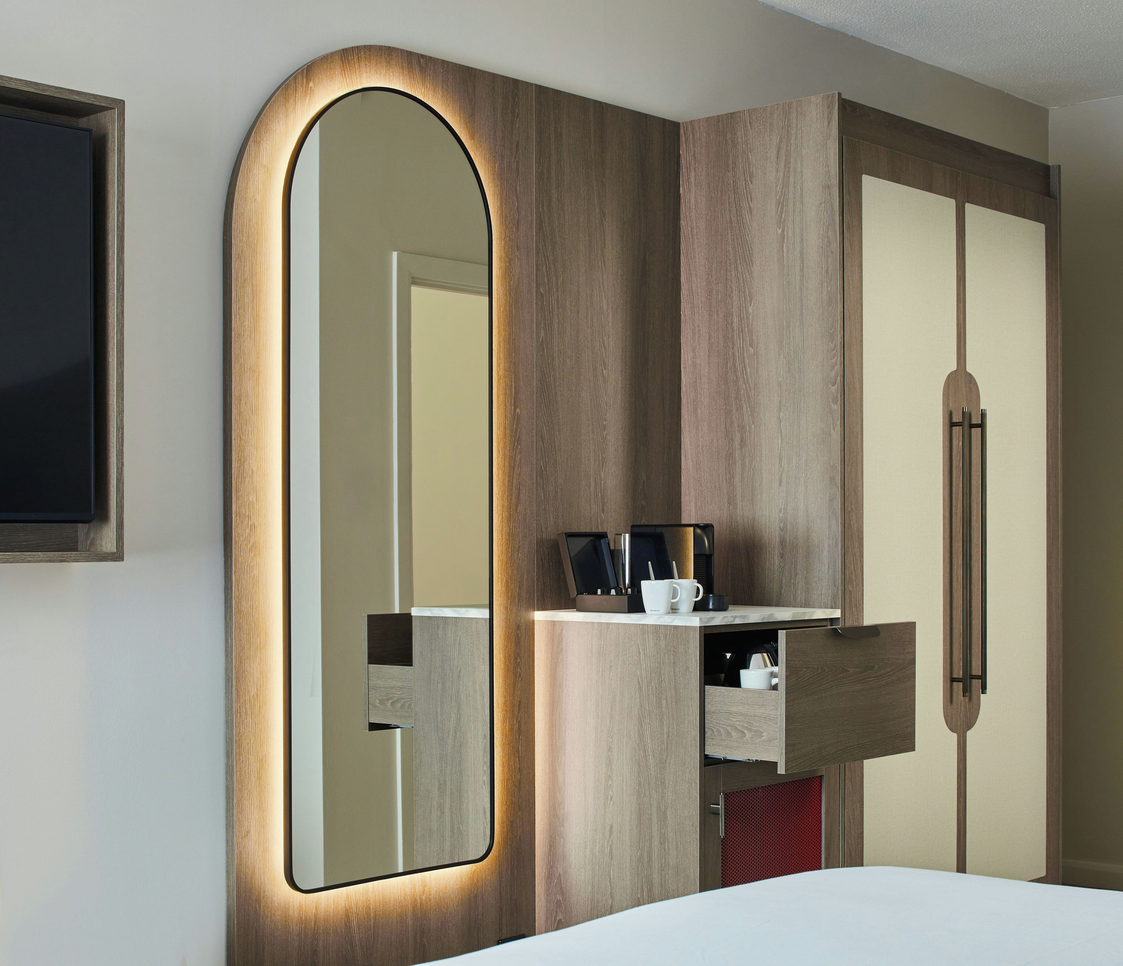 A hotel room with a large mirror, a wardrobe, and a small shelf with a coffee maker and mug.