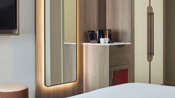 A hotel room with a large mirror, wooden wardrobe, and a small shelf with a coffee cup and utensils.