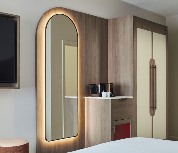 A hotel room with a large mirror, wooden wardrobe, and a small shelf with a coffee cup and utensils. - Leonardo Hotel Newcastle (Newcastle-upon-Tyne)