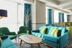 Un vestíbulo de hotel con un sofá azul verdoso, sillones verdes, una mesa de centro de madera y una planta decorativa.