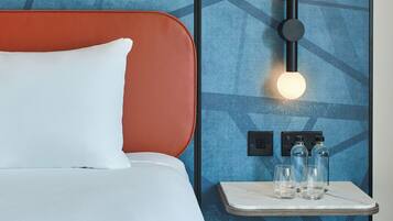 A hotel room with a bed, a nightstand with glasses and bottles, and a wall-mounted light fixture.