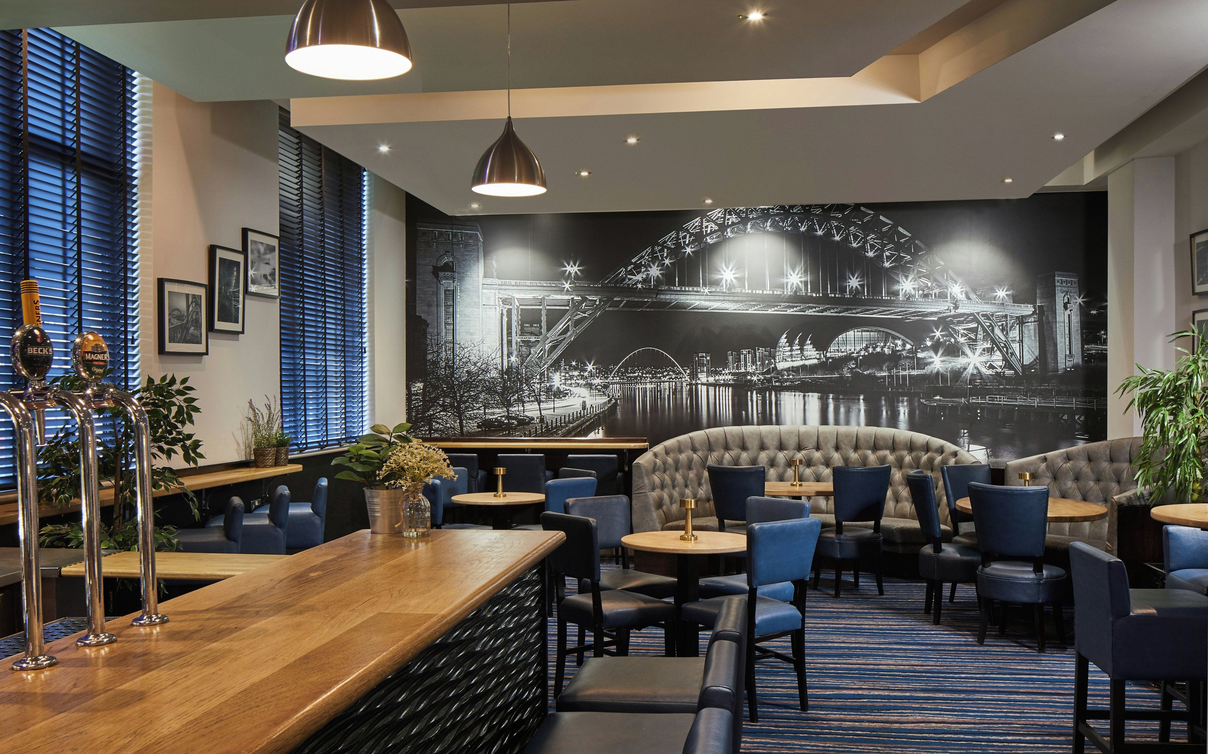 A modern bar area with a large mural of a bridge, seating with blue chairs, and a wooden bar counter.