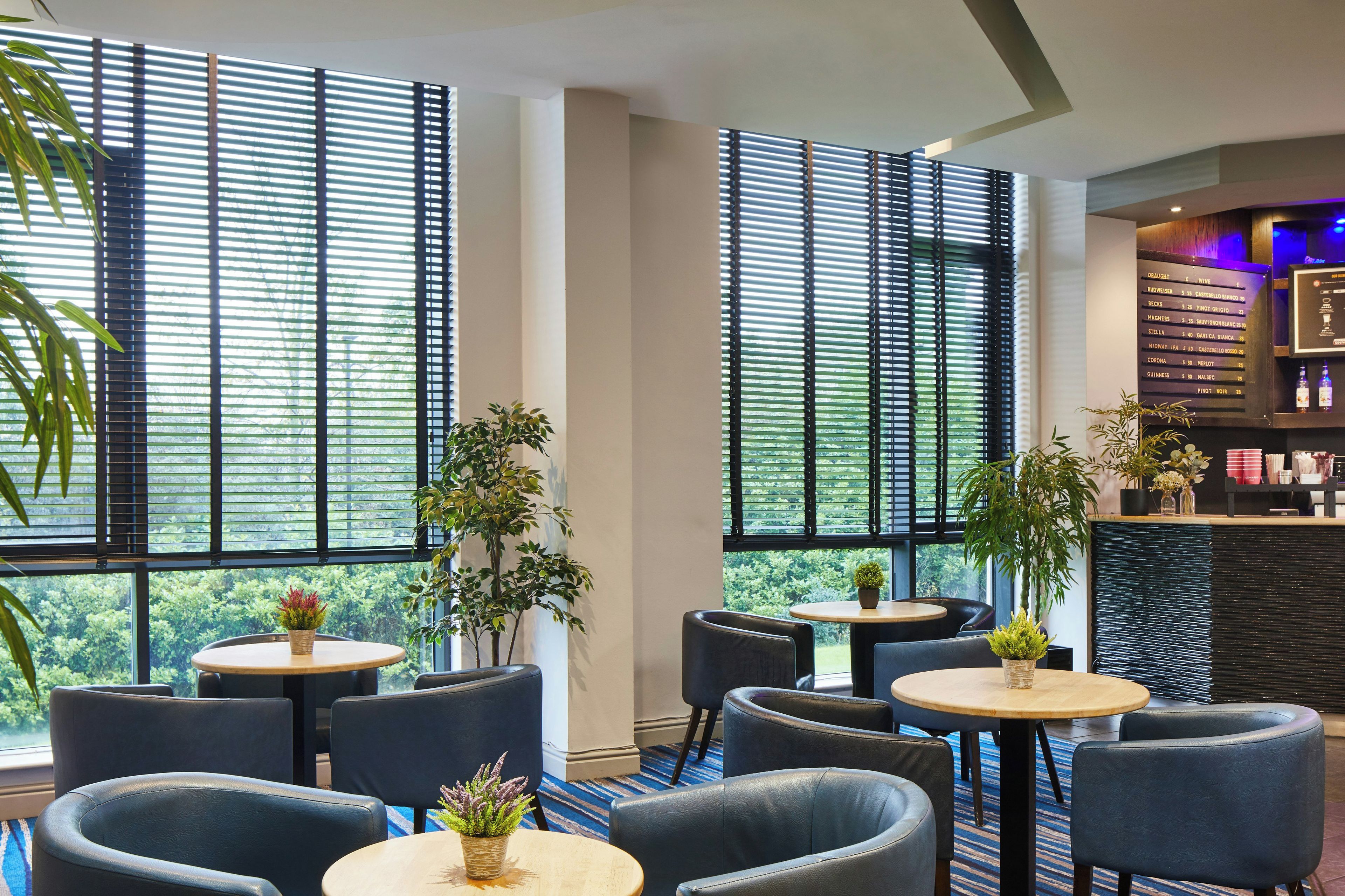 Un intérieur moderne avec des tables et des chaises rondes, de grandes fenêtres équipées de stores et des plantes d’intérieur.