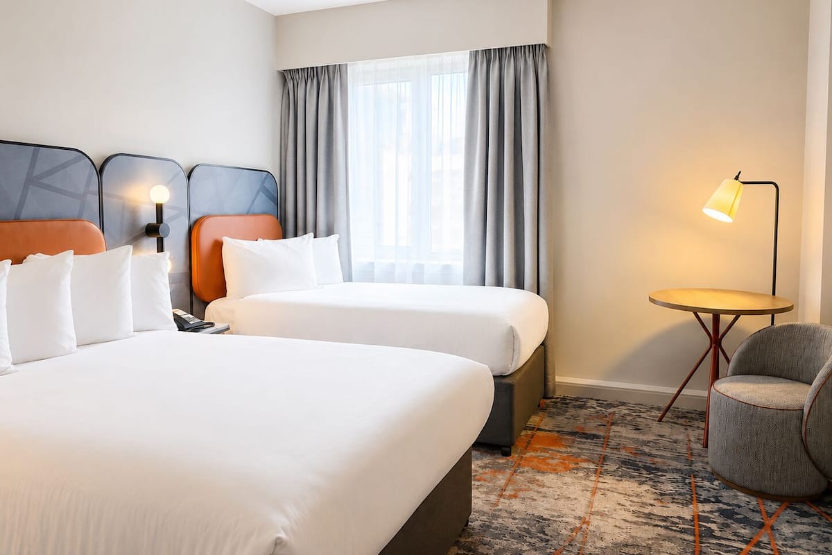 Une chambre d’hôtel avec deux lits, un bureau, une chaise, une lampe et une fenêtre avec des rideaux.
