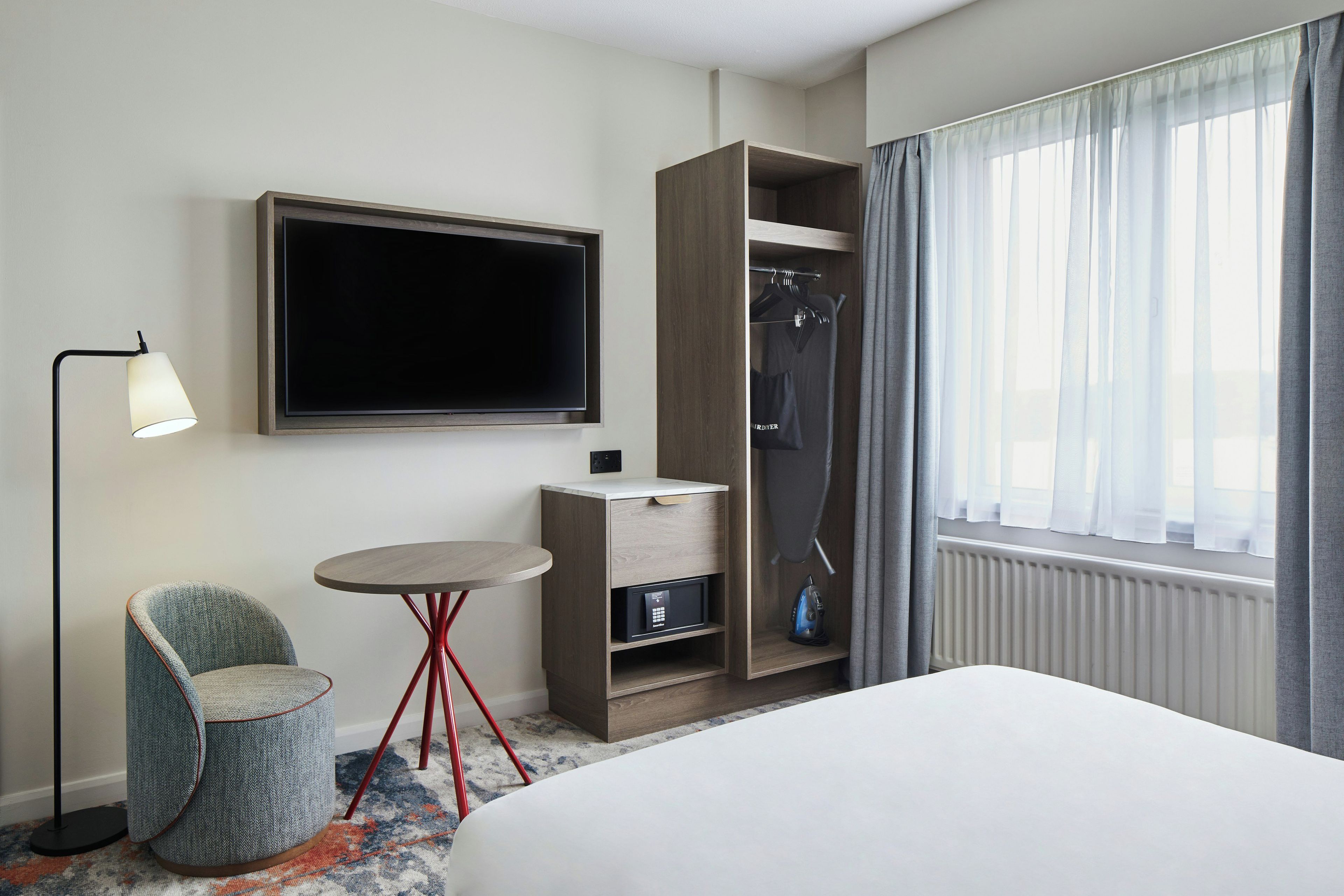 Een hotelkamer met een bed, een aan de muur bevestigde televisie, een kleine tafel, een stoel, een kledingkast en een raam met gordijnen.