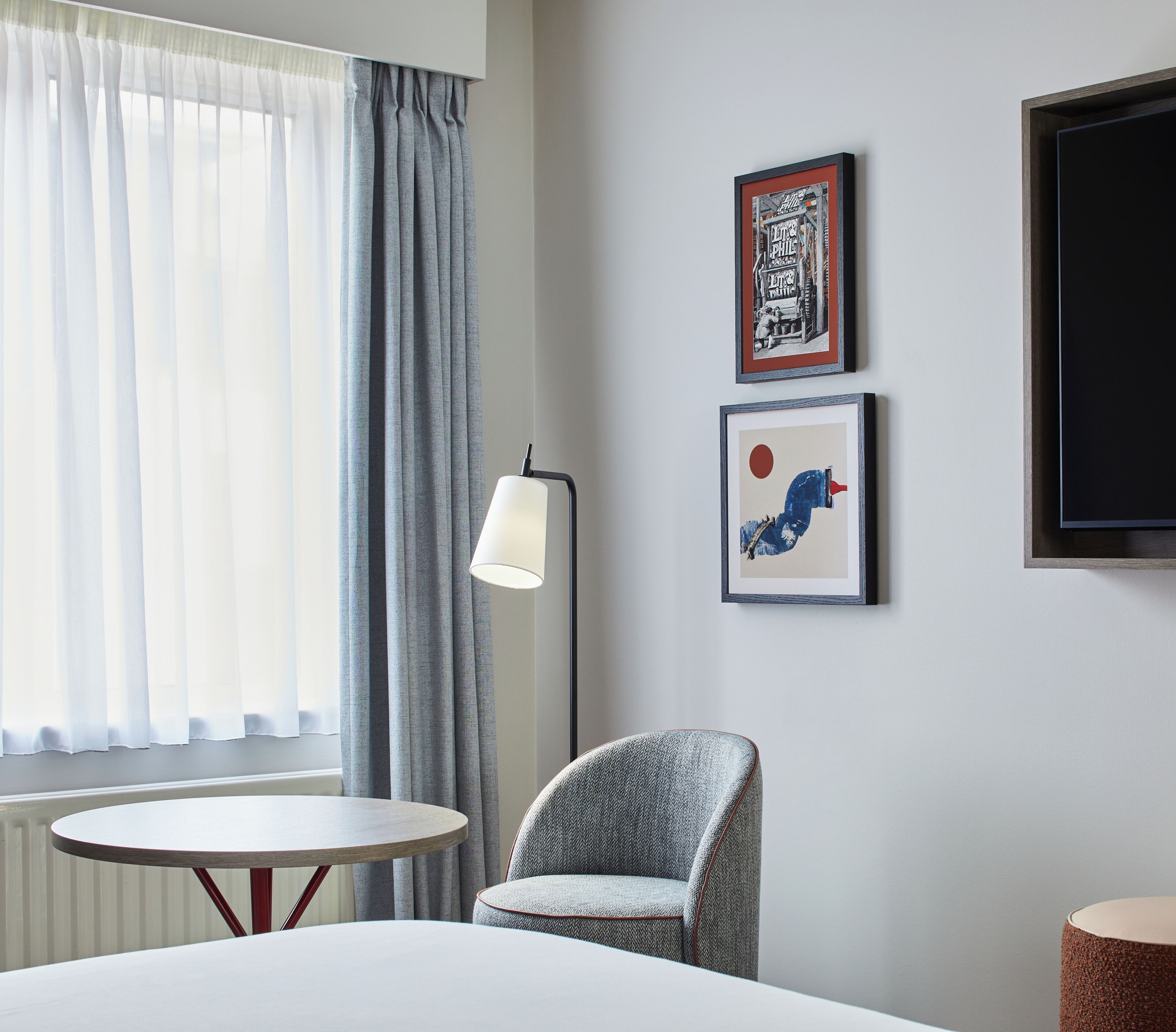 A hotel room with a bed, a round table, a chair, a floor lamp, and two framed pictures on the wall.