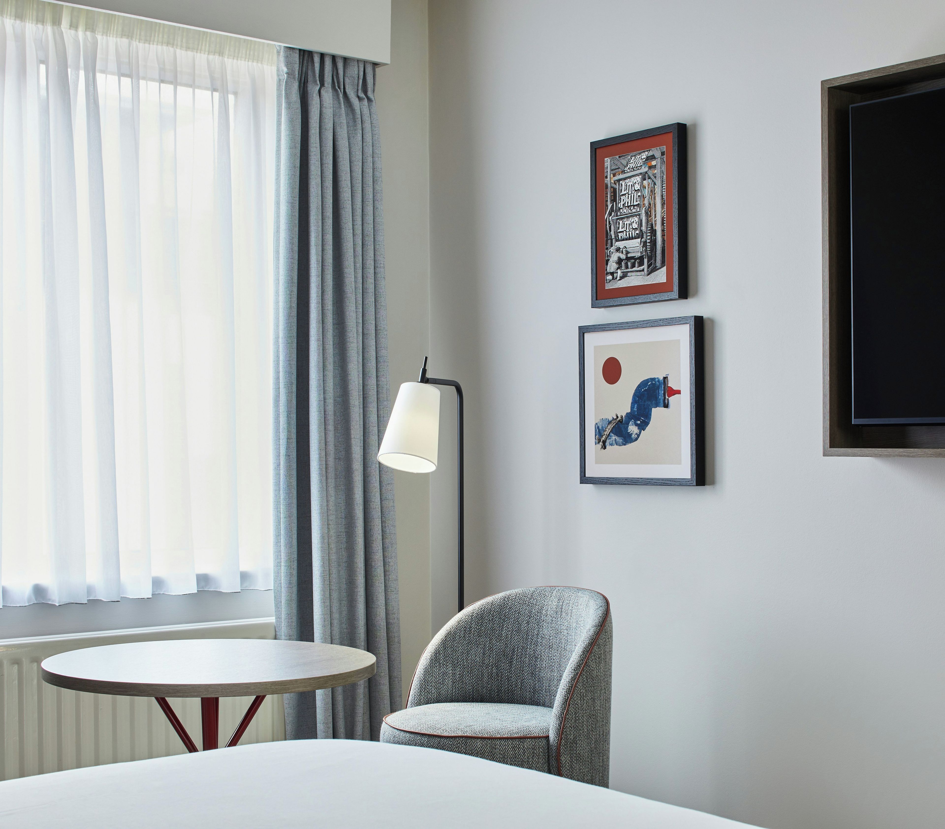 Una habitación de hotel con una cama, una mesa redonda, una silla, una lámpara de pie y dos cuadros en la pared.