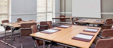 A conference room with multiple tables and chairs, a projector screen, and large windows with blinds.