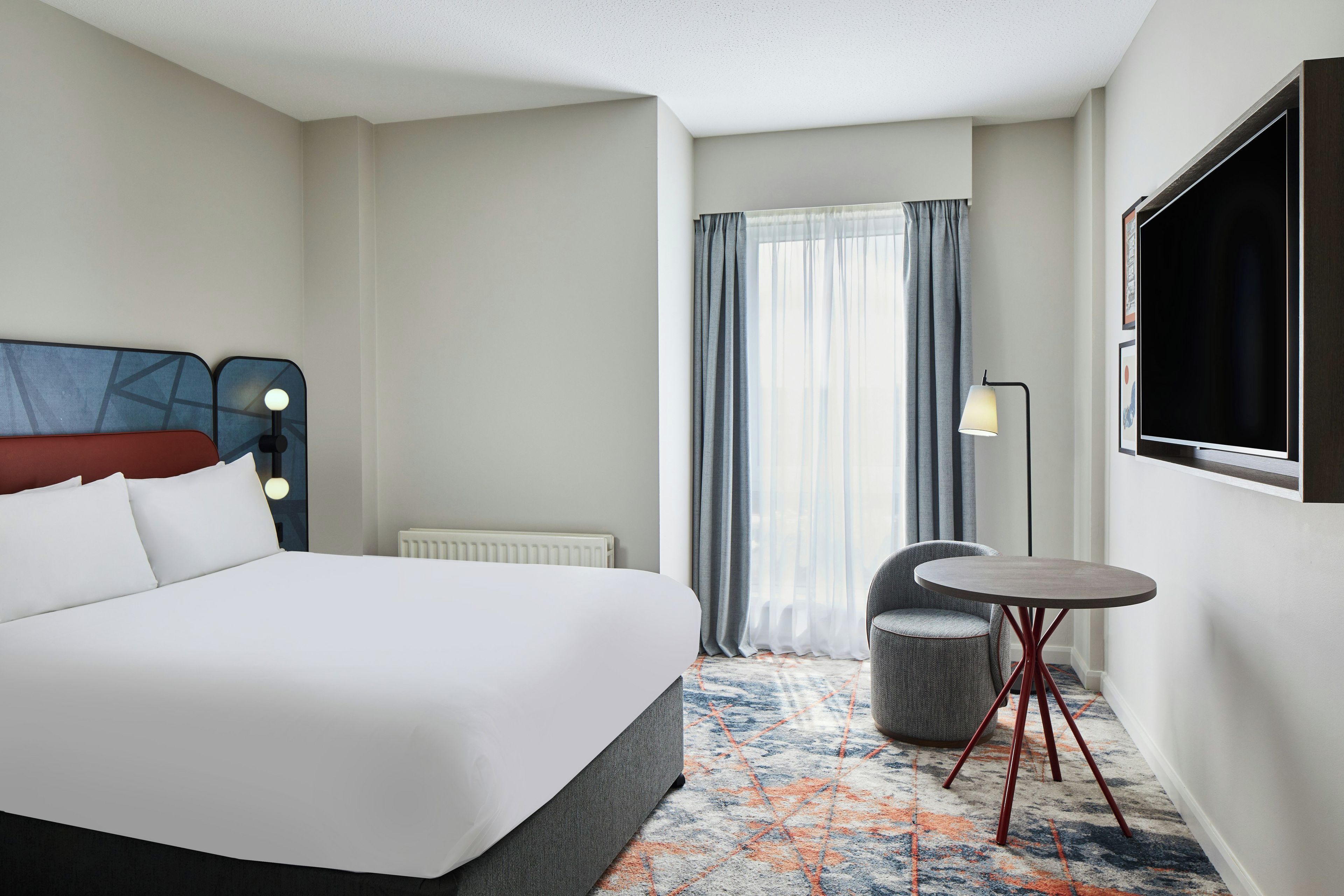 A hotel room with a bed, a chair, a small table, a TV, and a window with curtains.