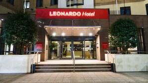 The exterior of the Leonardo Hotel with a prominent red sign, a set of stairs leading to the entrance, and two trees flanking the entrance.