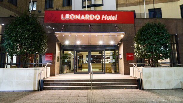 The exterior of the Leonardo Hotel with a prominent red sign, a set of stairs leading to the entrance, and two trees flanking the entrance. - Leonardo Hotel Newcastle (Newcastle-upon-Tyne)