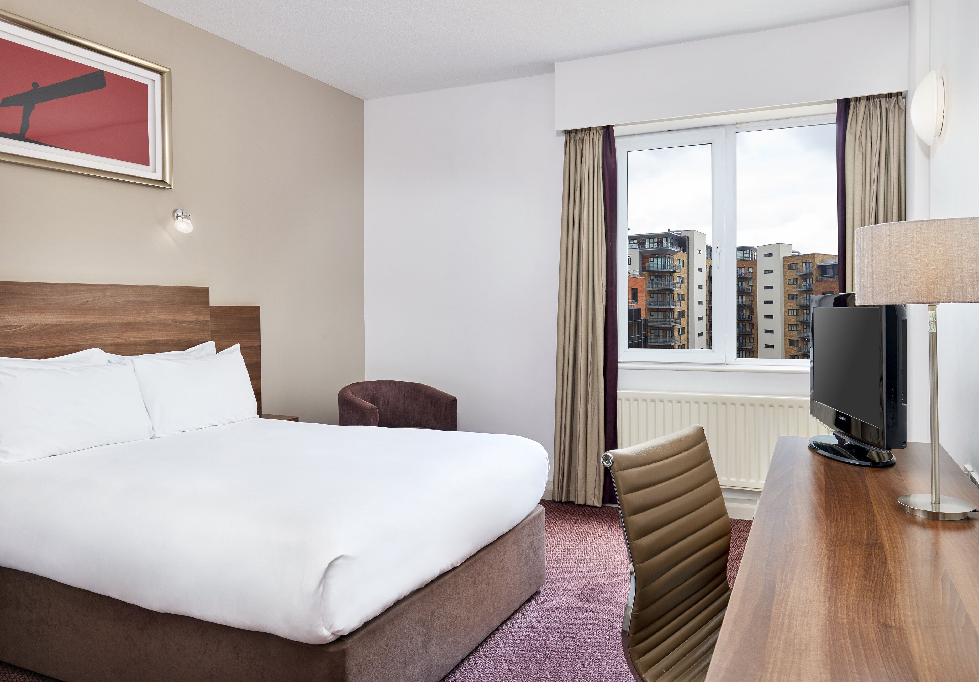 A hotel room with a bed, a desk with a television, a chair, and a window with curtains.