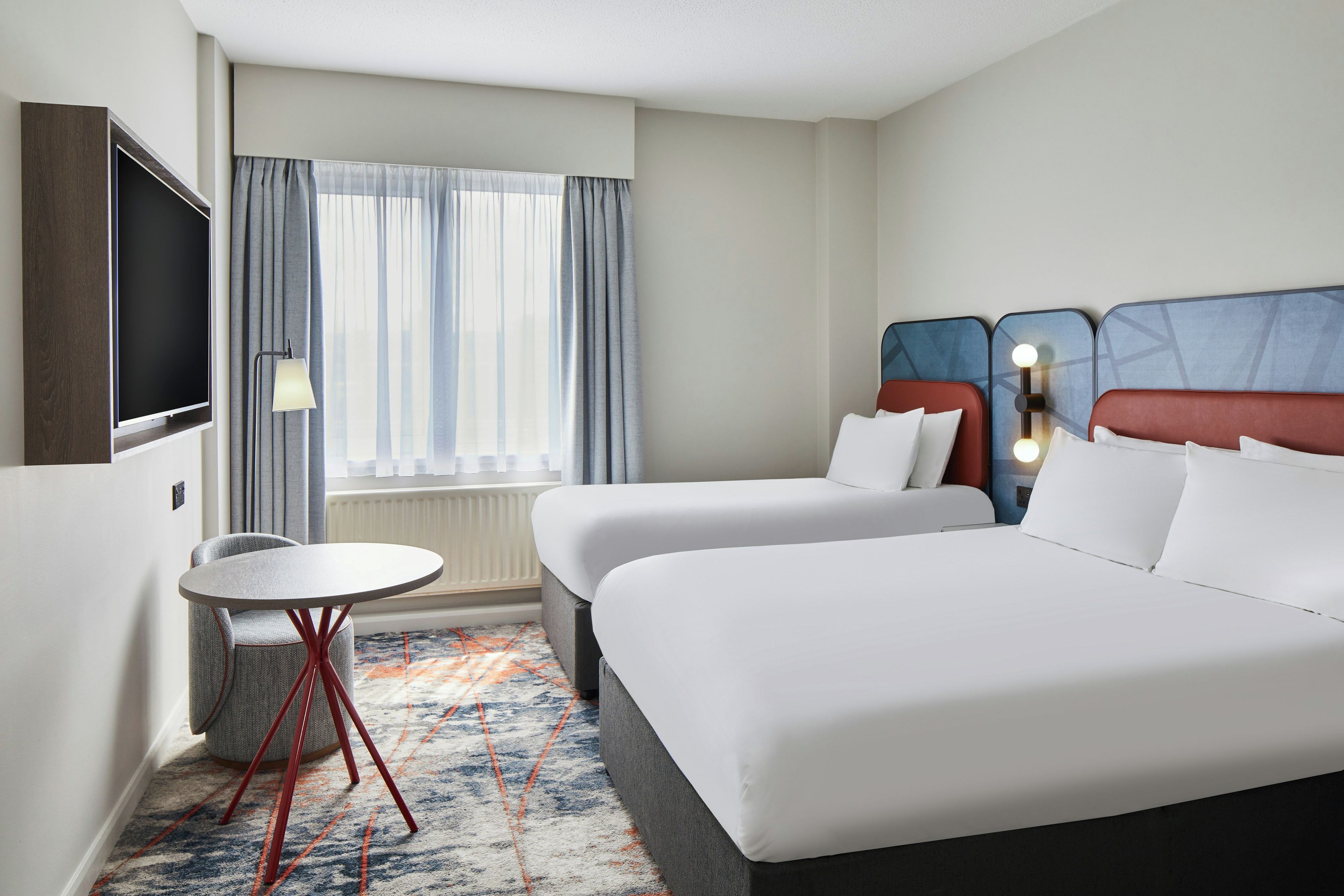 A hotel room with two beds, a TV, a small table, and a window with curtains.