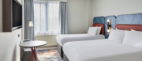 A hotel room with two beds, a TV, a small table, and a window with curtains.