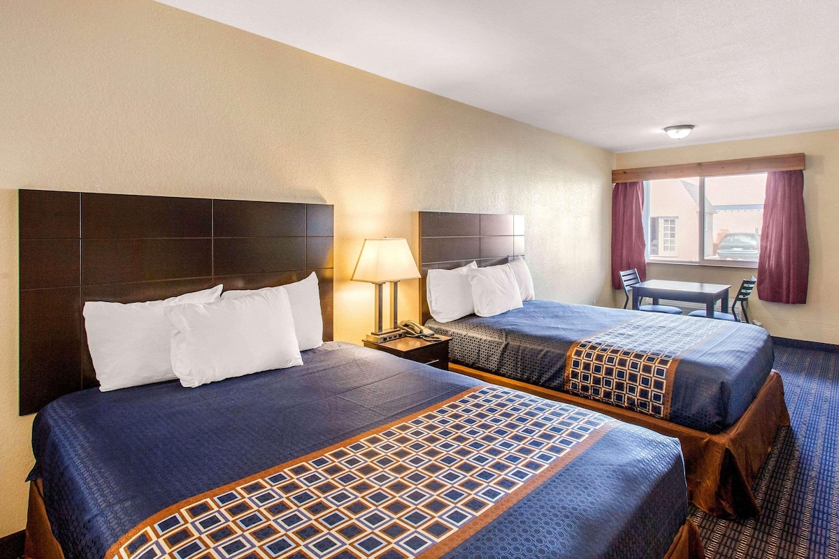 Quarto de hotel com duas camas, uma mesa de cabeceira, uma luminária, uma janela com cortinas e vista para o exterior.