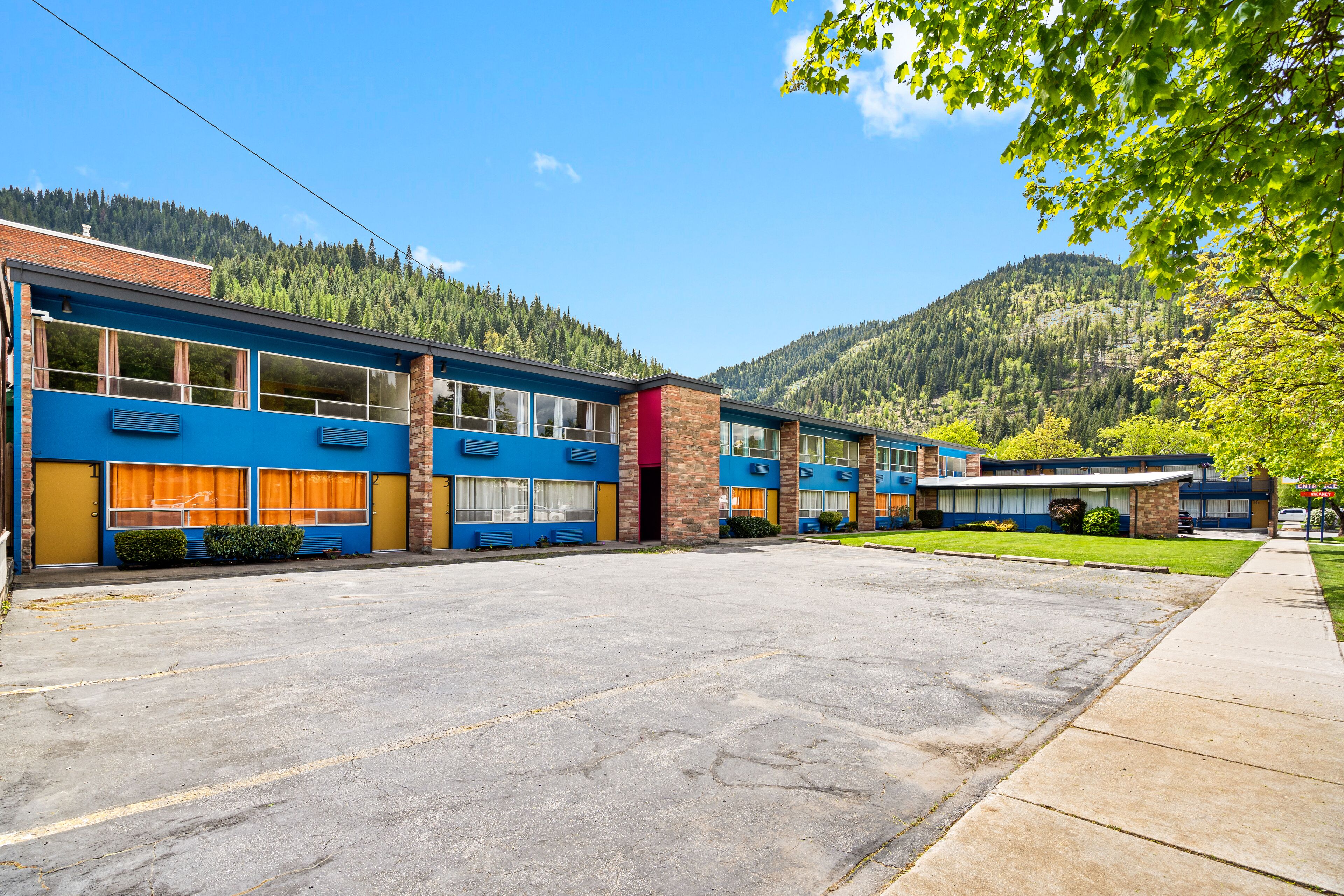 A row of blue and yellow motel buildings with large windows, a parking lot, and a mountain backdrop.