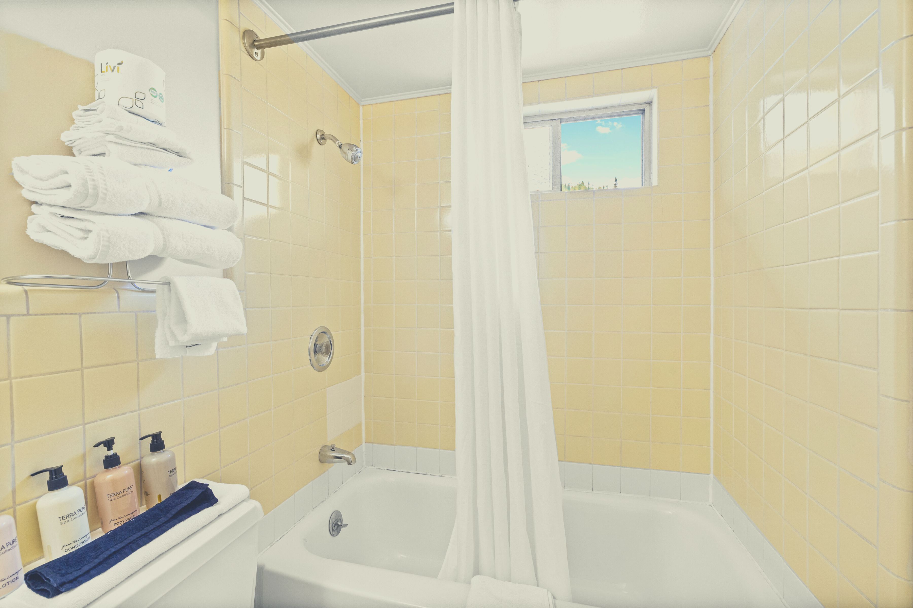 A bathroom with yellow tiled walls, a white bathtub, a shower area, and a window with a view.