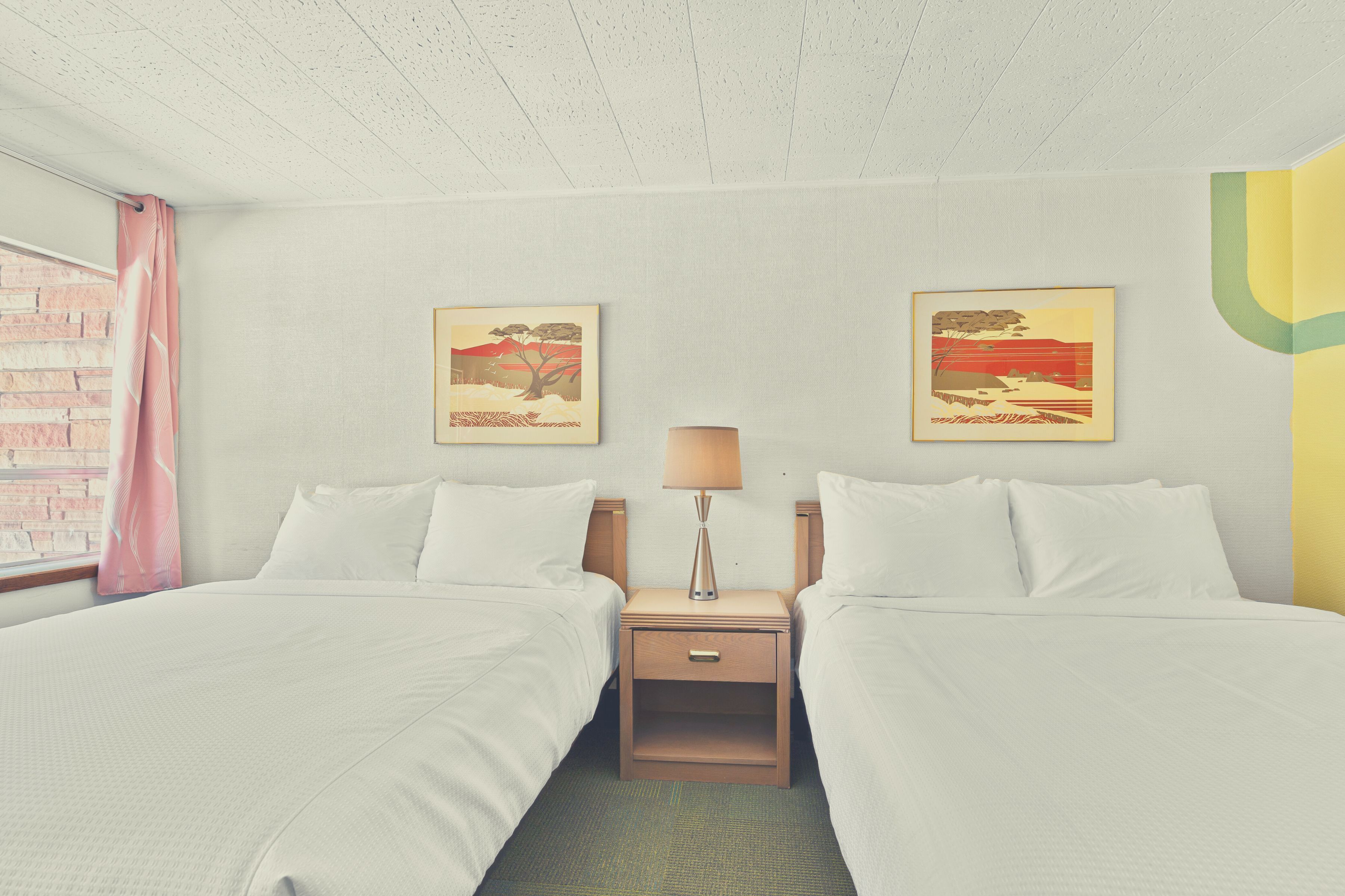 Habitación de hotel con dos camas, una mesita de noche con lámpara y dos cuadros colgados en la pared.