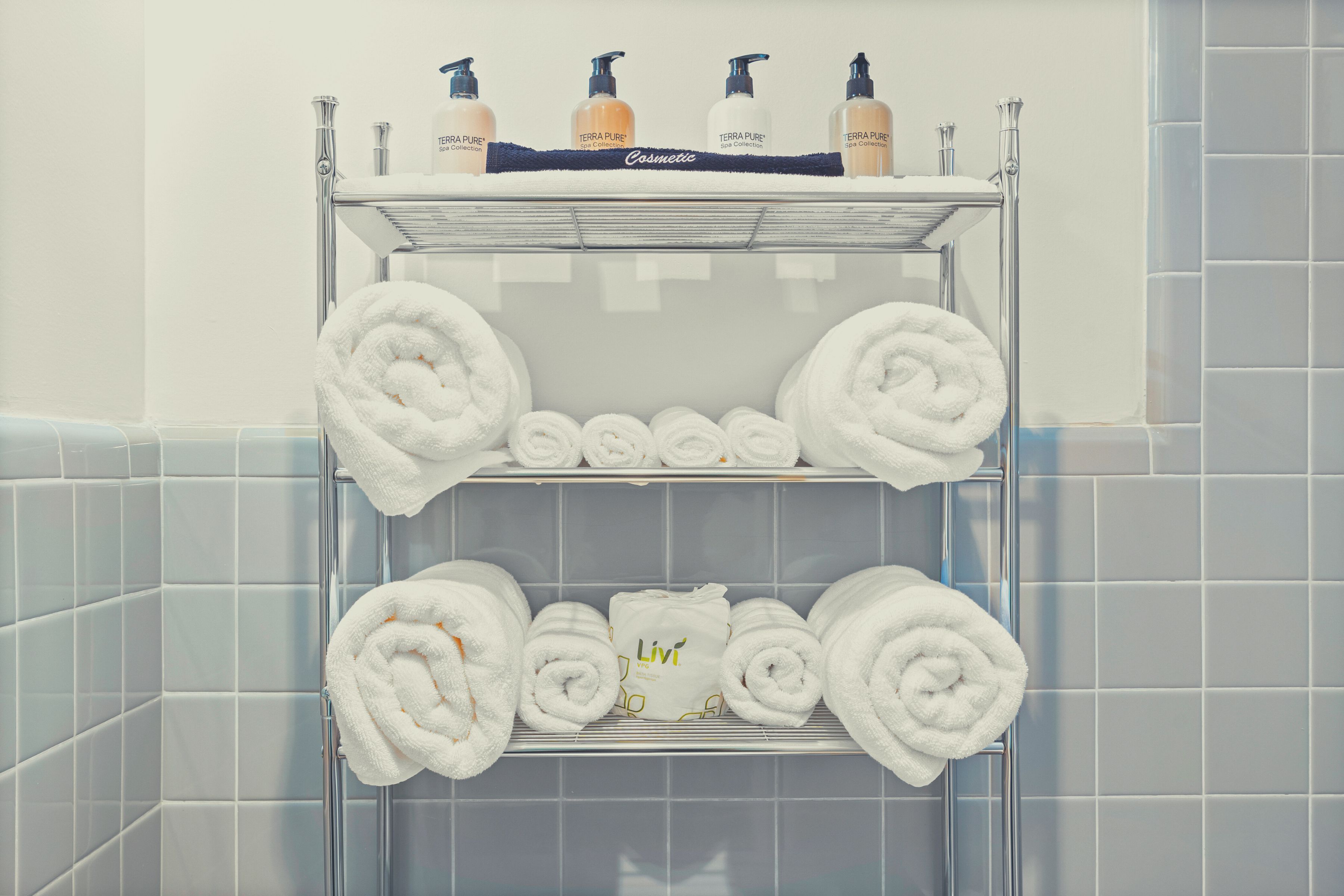 Een badkamer met een handdoekrek met meerdere witte handdoeken, een plank met Terra Pure toiletartikelen, en een Limi handdoek.