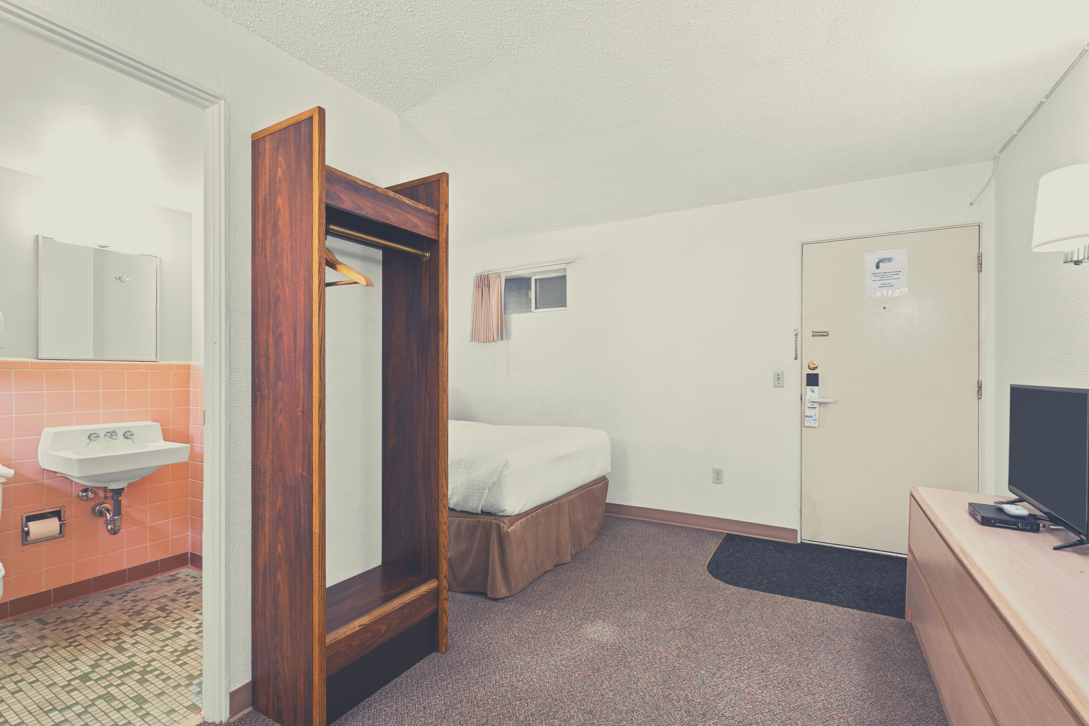 A hotel room with a wooden wardrobe, a single bed, a small window, a door with a sign, a sink, and a television on a dresser.